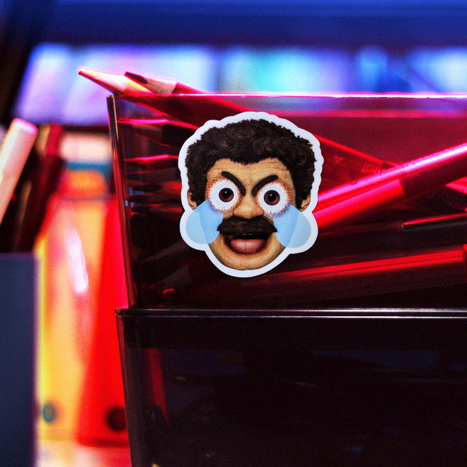 Kiszkiloszki sticker slapped on a plastic transparent container with crayons. A head of the AAAaaa Guy from Kiszkiloszki animation. The man is screaming in a mix of anger and sadness, with an open mouth and comic-like blue tears streaming from his wide open brown eyes.