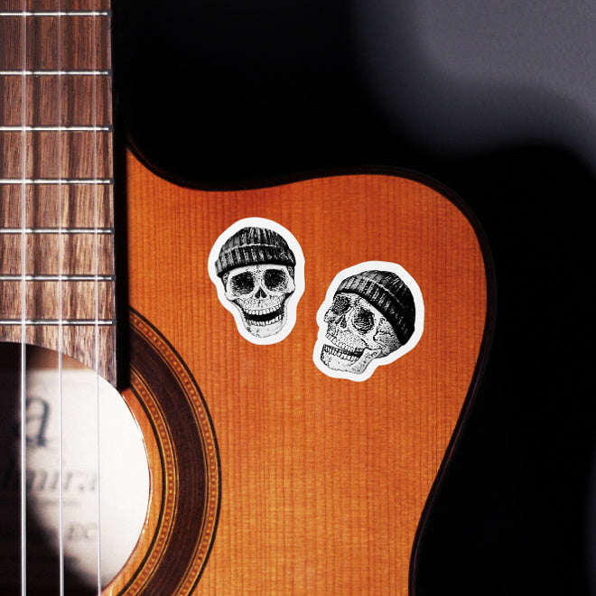 Kiszkiloszki stickers slapped on a front of a guitar. Two smiling skulls in black beanies.
