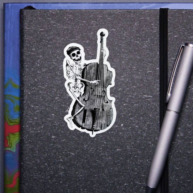 Kiszkiloszki sticker slapped on a gray notebook. A happy skeleton jazzman in a cap is playing on an old, broken double bass.