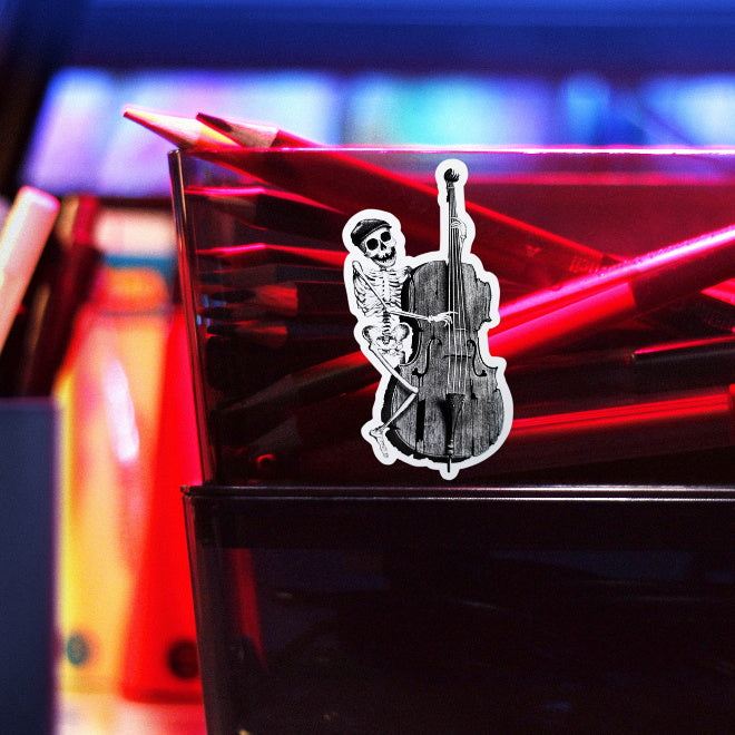 Kiszkiloszki sticker slapped on a plastic transparent container with crayons. A happy skeleton jazzman in a cap is playing on an old, broken double bass.