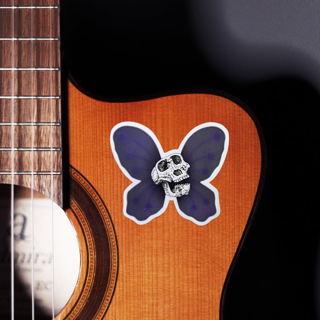 Kiszkiloszki sticker slapped on a front of a guitar. A Lucy skull smiling with a purple butterfly wings behind it. An alternative version of the Death Fairy Helper, character from the DFT universe.