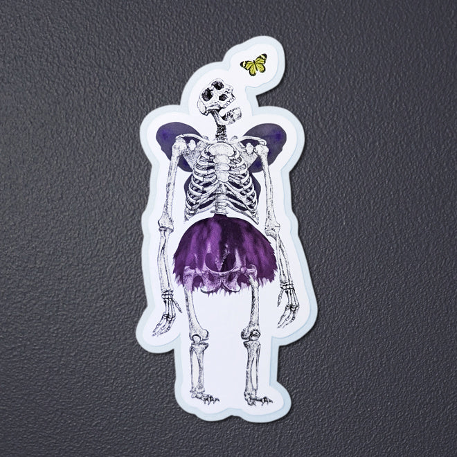 Kiszkiloszki sticker laying on a gray background. A massive skeleton in a comically small purple tutu and with purple butterfly wings is opening his mouth in an awe looking at a small butterfly flying close to his head.