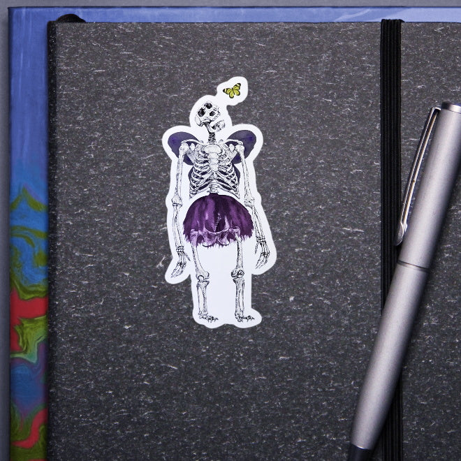 Kiszkiloszki sticker slapped on a gray notebook. A massive skeleton in a comically small purple tutu and with purple butterfly wings is opening his mouth in an awe looking at a small butterfly flying close to his head.