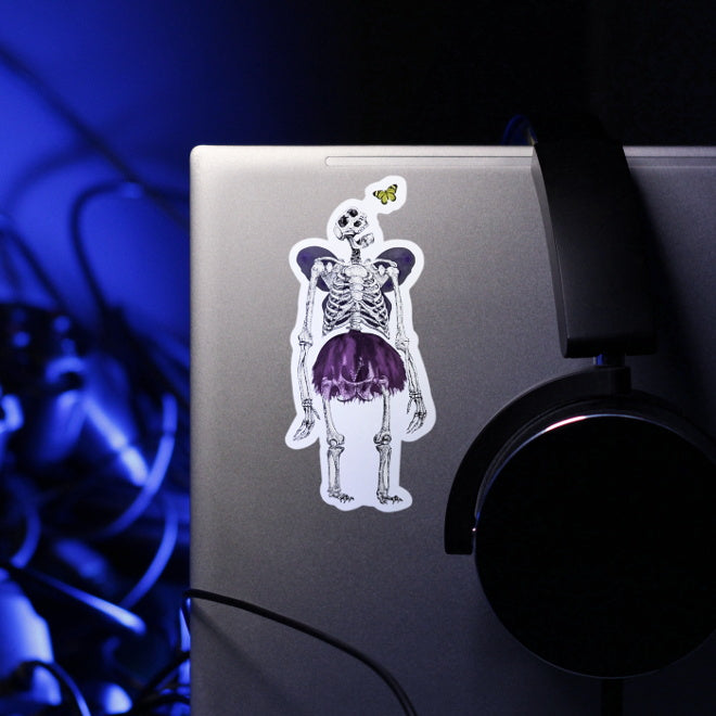 Kiszkiloszki sticker slapped on a silver laptop. A massive skeleton in a comically small purple tutu and with purple butterfly wings is opening his mouth in an awe looking at a small butterfly flying close to his head.