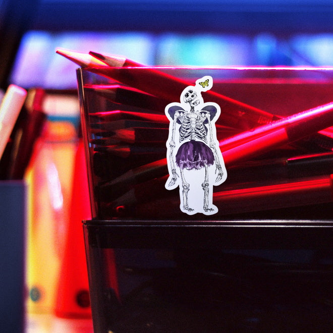 Kiszkiloszki sticker slapped on a plastic transparent container with crayons. A massive skeleton in a comically small purple tutu and with purple butterfly wings is opening his mouth in an awe looking at a small butterfly flying close to his head.