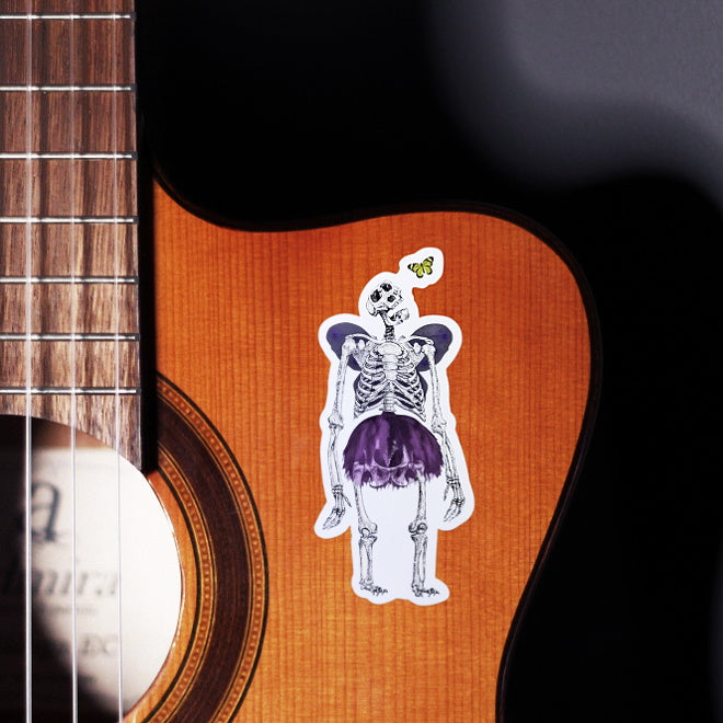 Kiszkiloszki sticker slapped on a front of a guitar. A massive skeleton in a comically small purple tutu and with purple butterfly wings is opening his mouth in an awe looking at a small butterfly flying close to his head.