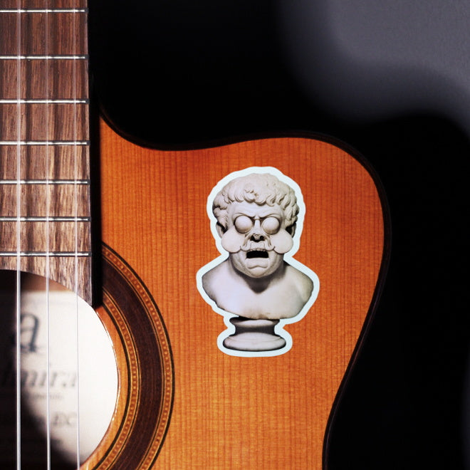 Kiszkiloszki sticker slapped on a front of a guitar. A bust sculpture of a furious mustache man with an open mouth in a scream and two comically sculpted tears.