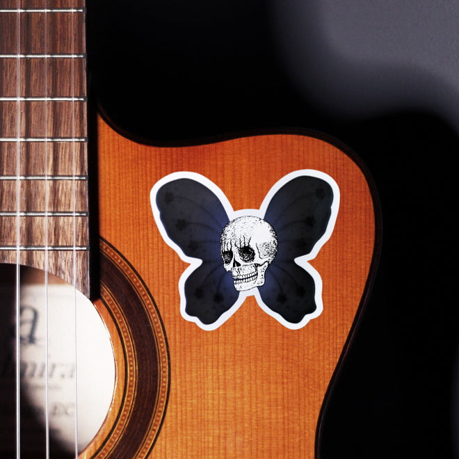 Kiszkiloszki sticker slapped on a front of a guitar. A maliciously smiling skull of the skeleton super hero with dark purple butterfly wings on the back.