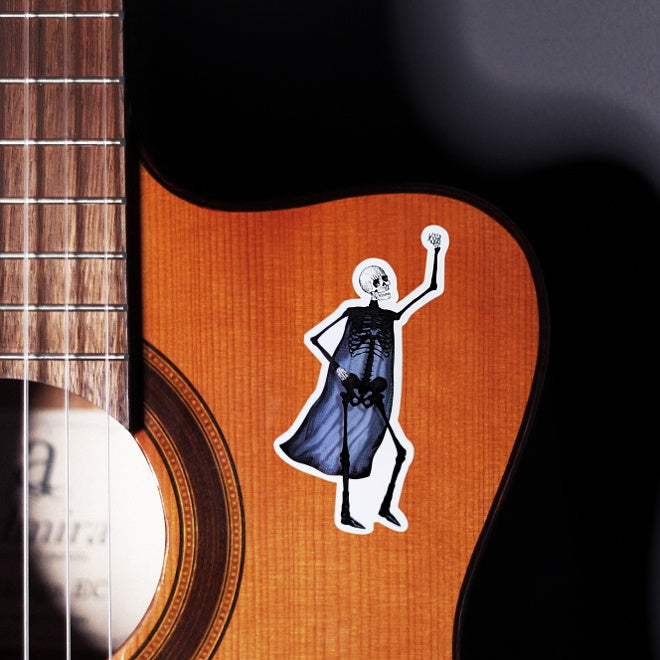 Kiszkiloszki sticker slapped on a front of a guitar. A happy skeleton super hero in a dark purple cape raises his hand in victory.