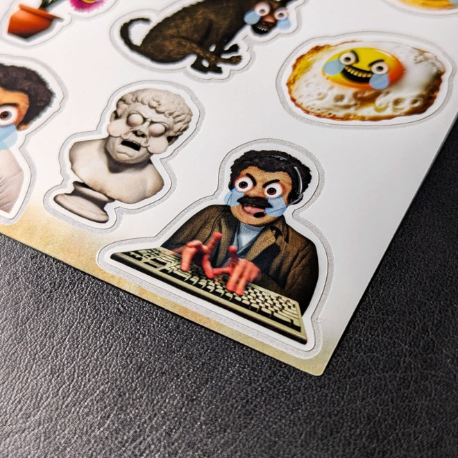 A close-up photo of a Kiszkiloszki sticker sheet placed on a table. The stickers include designs such as the AAAaaa Guy, an angry-sad man screaming while typing on a keyboard, with big blue tears running down his face, as well as an ancient bust, a dog, and a fried egg with similar features.