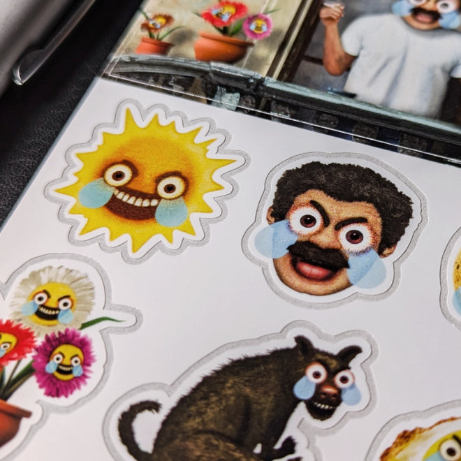 A close-up photo of a Kiszkiloszki sticker sheet placed on a table. The stickers include designs such as the AAAaaa Guy, an angry-sad man screaming, with big blue tears running down his face, as well as flowers, a sun, and a dog with similar features.