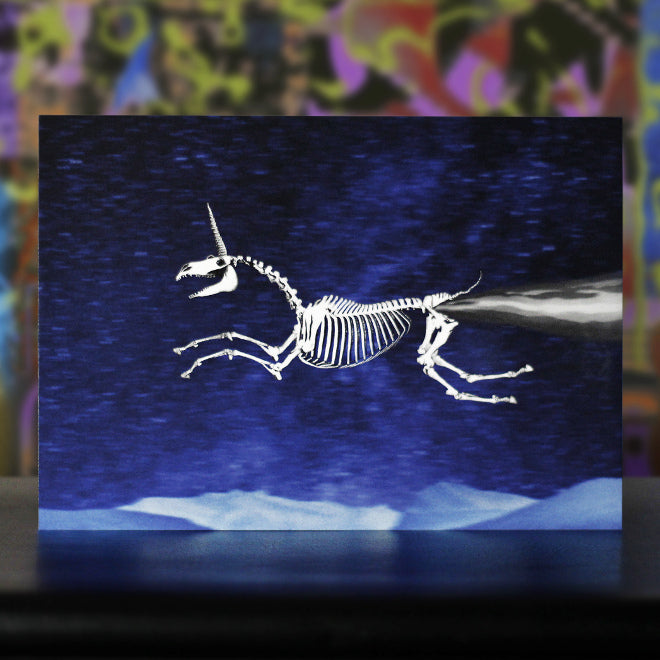 A close-up photo of a postcard featuring a flying skeletal unicorn with a wide smile and smoke comically jetting from its buttocks, set against a blurry, starry night sky.