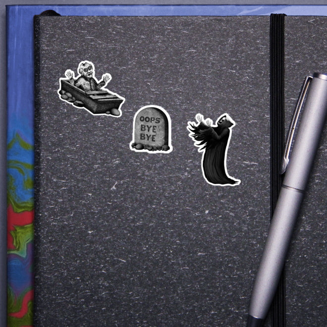 A visual representation of how Kiszkiloszki stickers would look on the cover of a notebook. Three monochrome stickers are displayed: a happy dead guy rising from a coffin, a gravestone inscribed with "Oops Bye Bye," and a smiling grim reaper with its arms raised, cloaked in flowing black. A silver pen clipped to the notebook completes the scene.