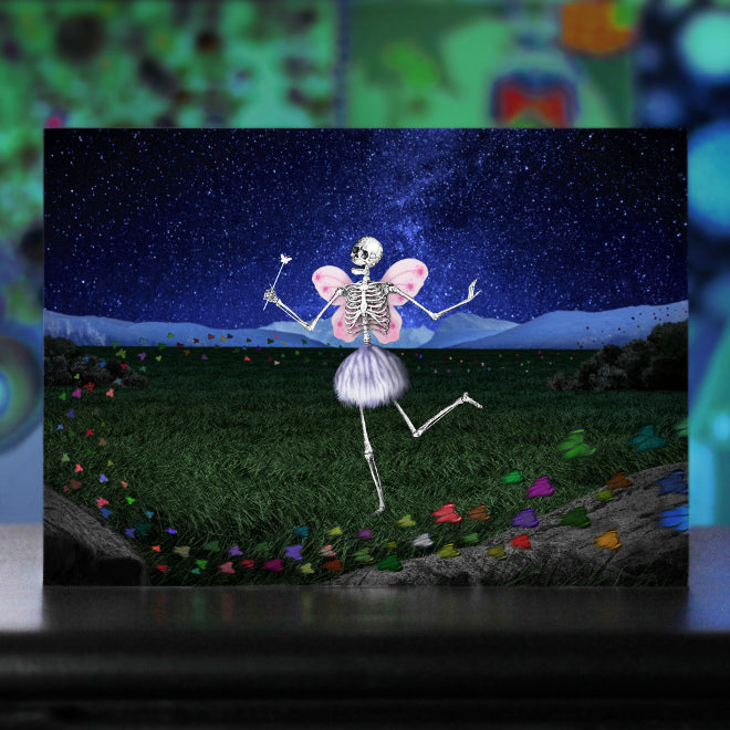 The Death Fairy. A close-up photo of an art postcard featuring a whimsical skeleton figure with butterfly-like pink wings and a small magical wand. The skeleton is positioned against a vibrant, starry blue background. Colorful butterflies are flying around the Death Fairy.