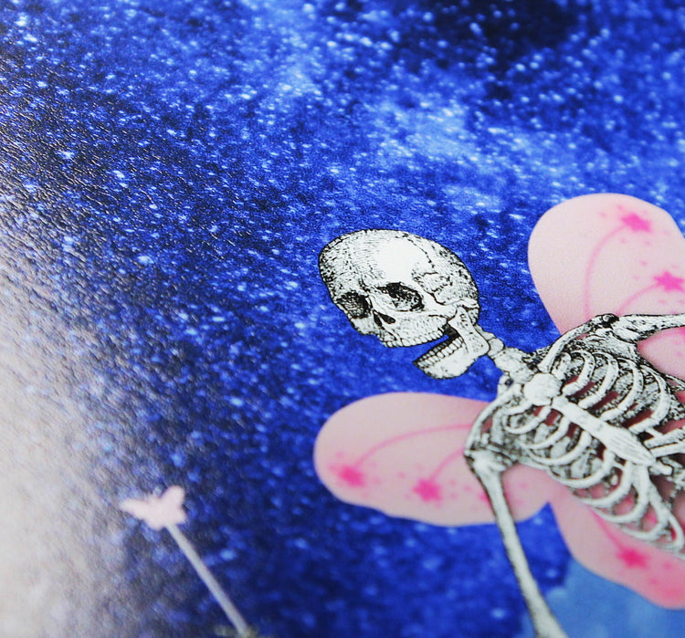 A close-up photo of an art print featuring a whimsical skeleton figure with butterfly-like pink wings and a small magical wand. The skeleton is positioned against a vibrant, starry blue background. 