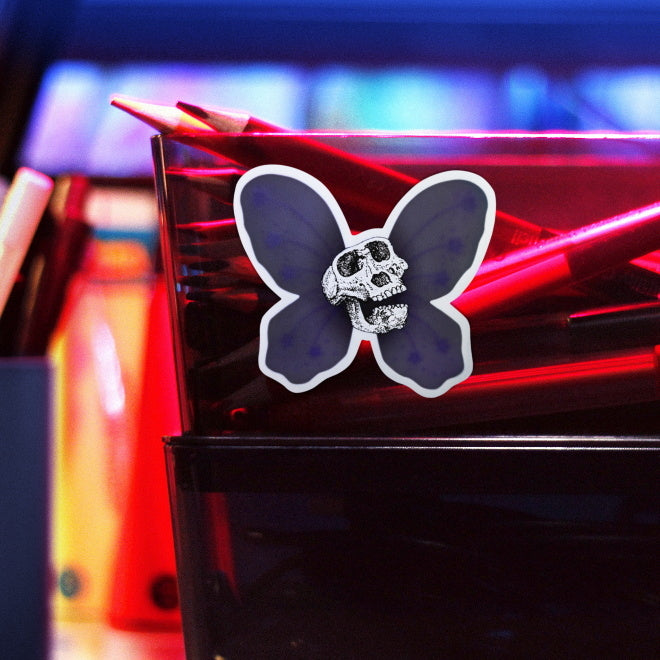 Kiszkiloszki sticker slapped on a plastic transparent container with crayons. A Lucy skull smiling with a purple butterfly wings behind it. An alternative version of the Death Fairy Helper, character from the DFT universe.