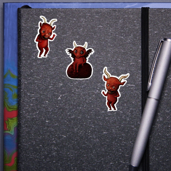 A visual representation of how Kiszkiloszki stickers would look on the cover of a notebook. Three stickers featuring mischievous, red devil-like creatures with horns. The designs include two standing devils in playful poses and one devil perched on a rock with bat-like wings. A silver pen clipped to the notebook completes the scene.