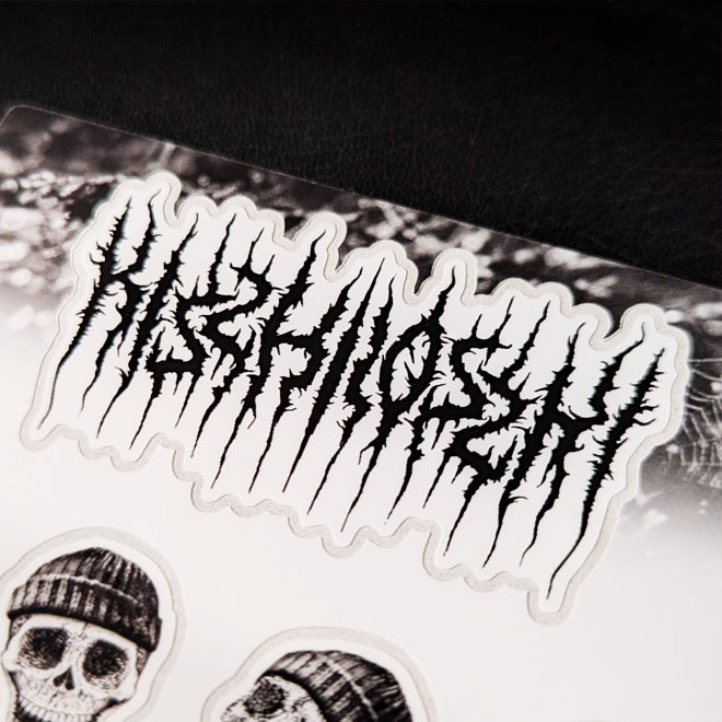 A close-up photo of a Kiszkiloszki sticker sheet placed on a table. The stickers include designs such as smiling skulls wearing beanies and the Kiszkiloszki logo designed in the style of a metal band.