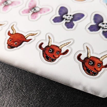 A close-up photo of a Kiszkiloszki sticker sheet placed on a table. The stickers include designs such as smiling red devils with horns and butterfly skulls from Kiszkiloszki animations.