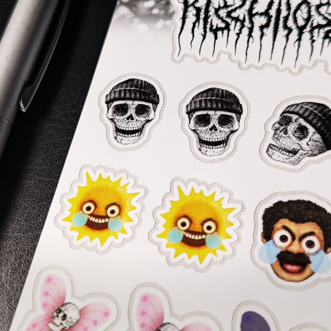 A close-up photo of a Kiszkiloszki sticker sheet placed on a table. The stickers include designs such as smiling skulls wearing beanies, an angry-sad sun with cartoonish blue tears, and an angry-sad face of a man with wide eyes and large blue tears.