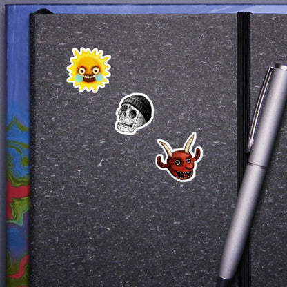 A visual showcase of Kiszkiloszki stickers on a gray notebook cover. Featured are three unique designs: an angry, crying sun; a smiling skull wearing a beanie; and the head of a happy red devil with horns. A silver pen clipped to the notebook completes the scene.