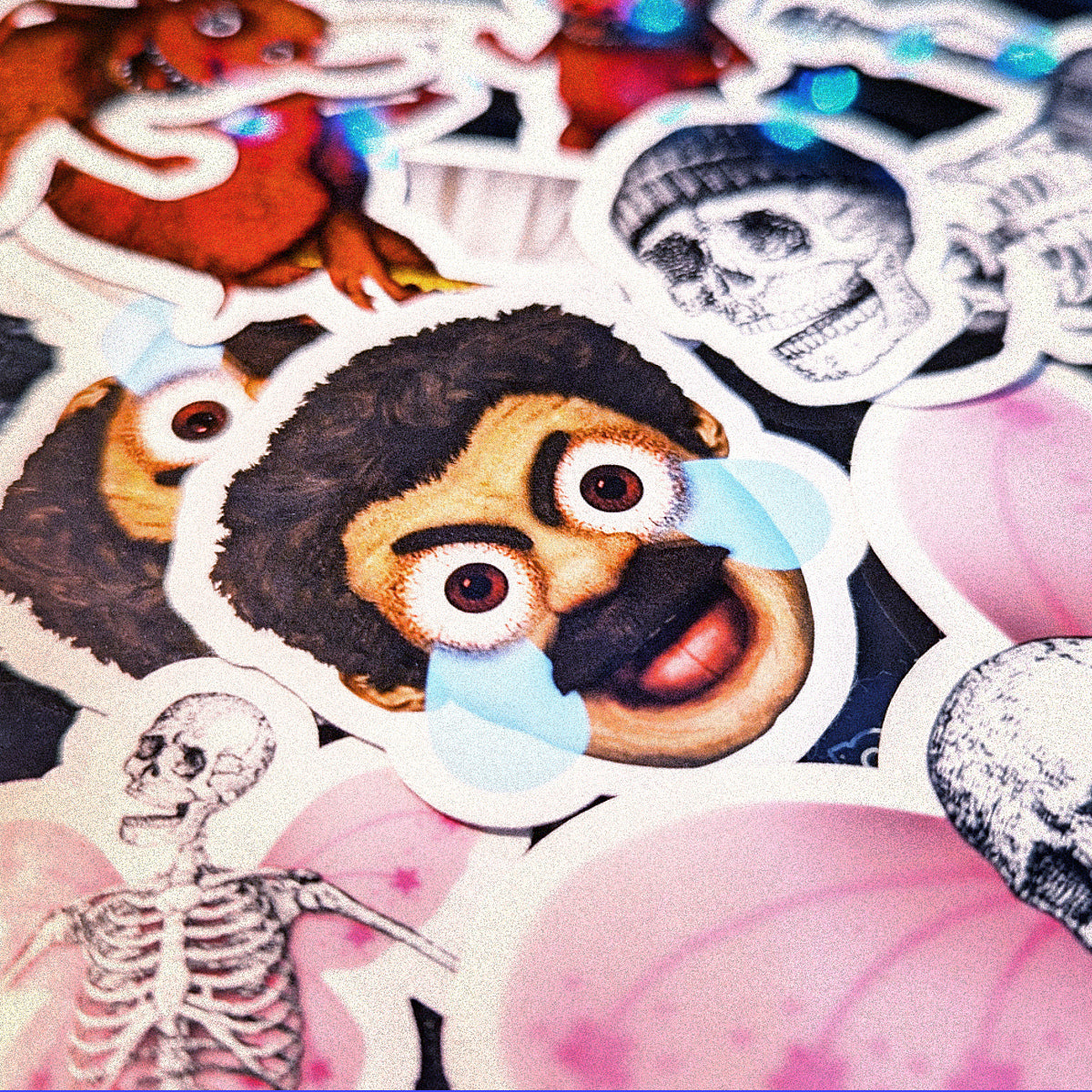 Stickers are chaotically piled on top of each other, with the frustrated face of a man with a mustache in the foreground, comically screaming and crying in anger. Stickers of cute red devils and smiling skeletons blur in the background.