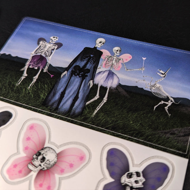 A close-up photo of a Kiszkiloszki sticker sheet placed on a table. The stickers include characters from Kiszkiloszki animations. The Death Fairy, The Oopsie-Man and other skeletal characters.