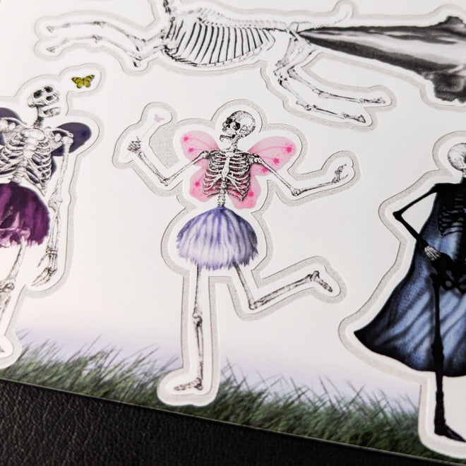 A close-up photo of a Kiszkiloszki sticker sheet placed on a table. The stickers include designs such as the Death Fairy and other skeletal characters from Kiszkiloszki's animations.