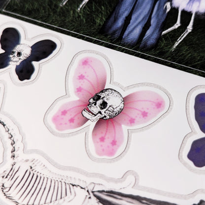 A close-up photo of a Kiszkiloszki sticker sheet placed on a table. The stickers include designs such as smiling Death Fairy Skull with pink butterfly wings.