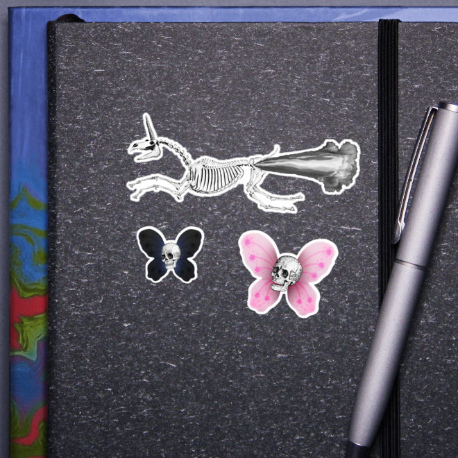 A visual representation of how Kiszkiloszki stickers would look on the cover of a notebook. The gray notebook features three unique stickers: a skeletal unicorn with smoke comically jetting from its buttocks as it flies, and two smaller butterfly-shaped stickers. Dark blue/purple one with a skull and one pink with a skull. A silver pen clipped to the notebook completes the scene.