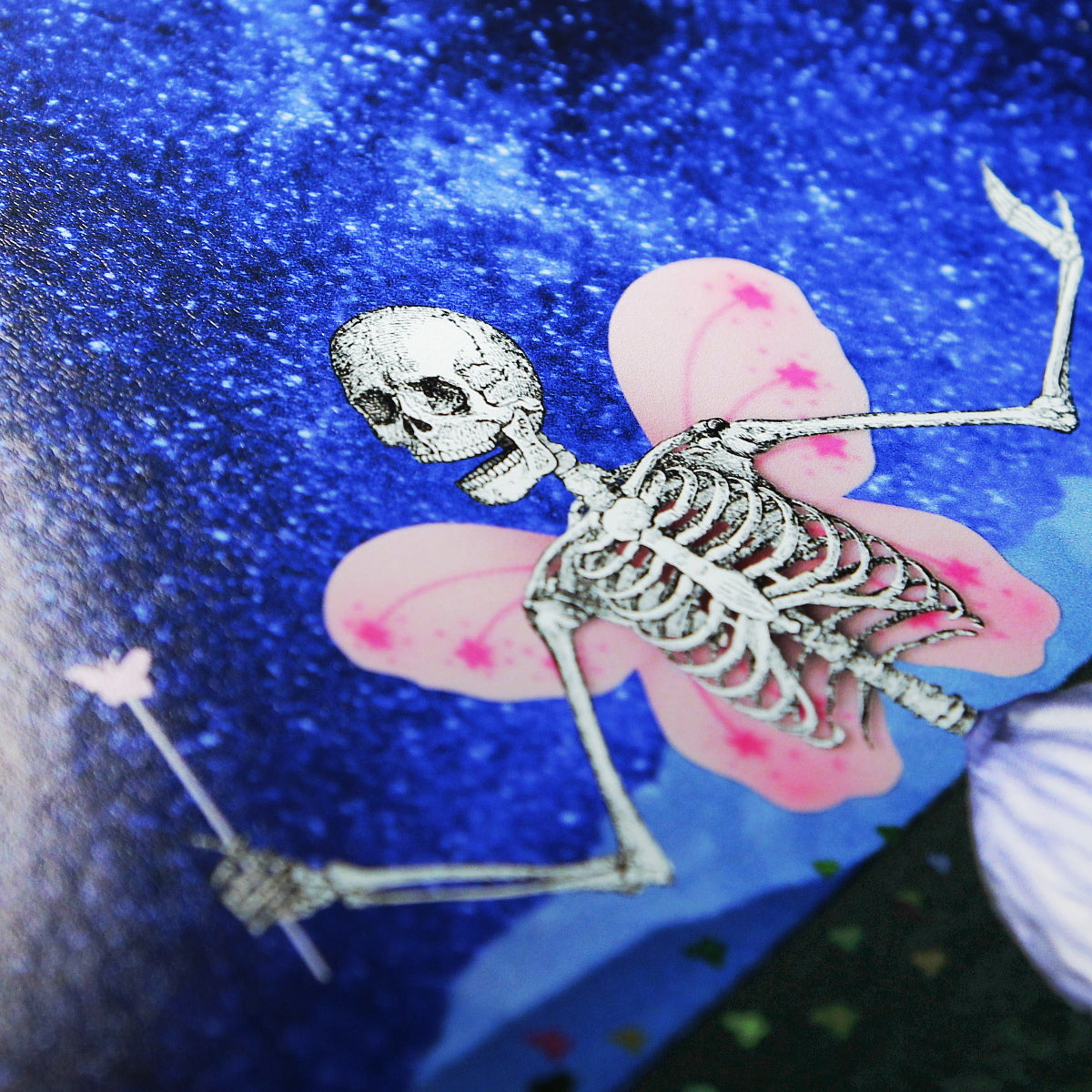 The Death Fairy. A close-up photo of an art print featuring a whimsical skeleton figure with butterfly-like pink wings and a small magical wand. The skeleton is positioned against a vibrant, starry blue background.