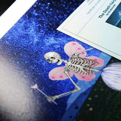 A close-up photo of an art print featuring a whimsical skeleton figure with butterfly-like pink wings and a small magical wand. The skeleton is positioned against a vibrant, starry blue background. Partially visible on the side is a "Certificate of Authenticity" for the artwork titled "The Death Fairy Tales" by Kiszkiloszki. The print has a glossy finish, and the details of the illustration, such as the wings and the skeletal structure, are finely detailed.