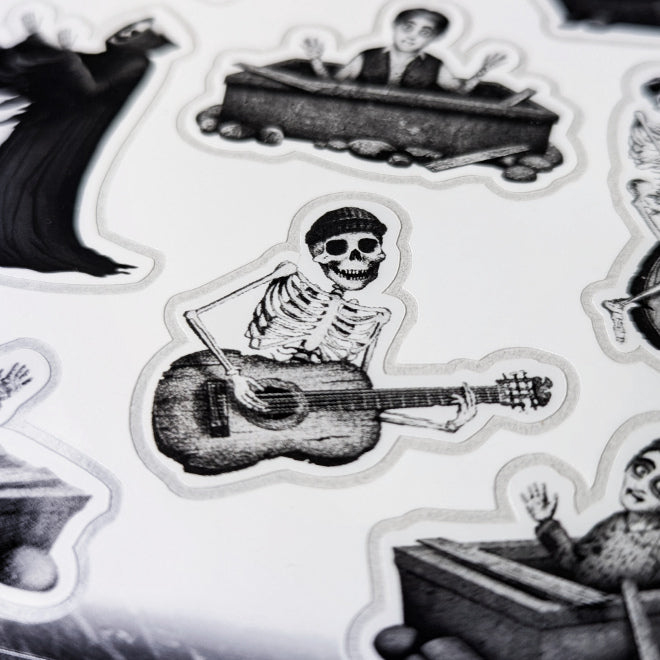 A close-up photo of a Kiszkiloszki sticker sheet. The stickers include designs such as happy peaople in their graves, dancing Death in a black robe, smiling skeleton playing guitar.